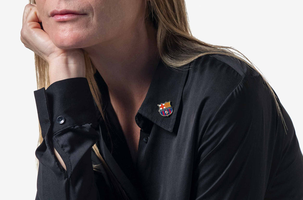Modelo mujer de pelo rubio y piel blanca con una camisa negra vistiendo la insignia del FC Barcelona acabada en oro de 18K y con un diamante engastado en su centro.